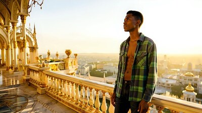 Standing on a palace balcony, a man in an unbuttoned plaid shirt looks out at sunset