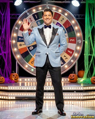 Smiling on a Halloween stage, a man in a light blue tuxedo waves near a game wheel