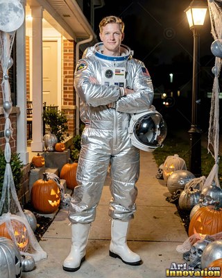 Standing on a front walkway, a happy man is wearing a silver astronaut suit near pumpkins