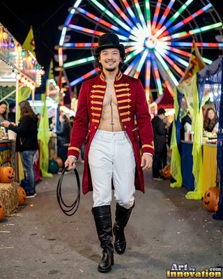 A man walks forward in an open red coat under colorful Ferris wheel lights