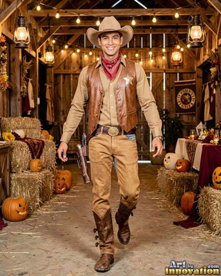 Walking forward in a rustic barn, a smiling man is dressed in a tan cowboy outfit