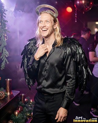 Smiling at a party, a blond man is dressed in black feathered wings, a dark satin shirt, and a halo