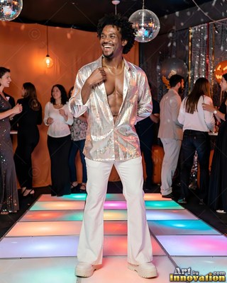 A smiling man dances in an unbuttoned silver shirt and white flares