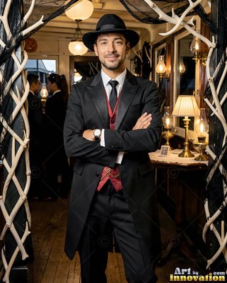 A man in a black fedora crosses his arms while wearing a dark suit with a red vest in warm lamplight