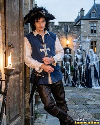 Leaning by a stone doorway, a man in a blue musketeer vest rests his sword
