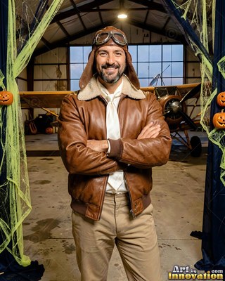 A smiling man in a brown bomber jacket crosses his arms in a warm yellow hangar light