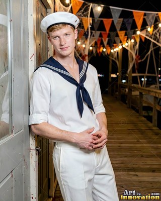 Leaning by a door, a blond man poses in a white sailor costume on a wooden pier