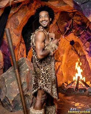 Standing in profile inside a cave, a cheerful man is wearing a leopard tunic by a fire