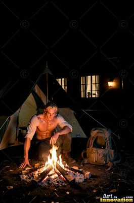 At a dark campsite, a blond man crouches by a campfire outside his tent