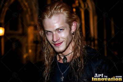 At night outside a church, a blond man smiles with pale eyes and black necklaces