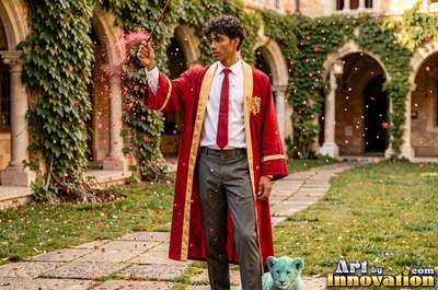 Standing in a castle courtyard, a young man raises a wand in a red robe