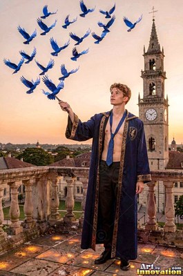 In a clear blue robe, a bare-chested man releases blue birds from his wand on a castle rooftop
