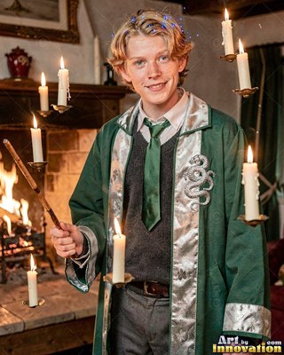 Holding a wooden wand, a young blond man in a green gown stands by floating candles