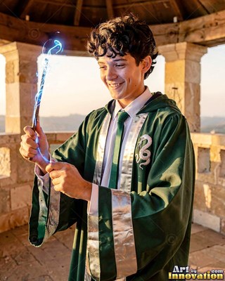 A smiling young man in a green gown holds a wand that sparks with blue light