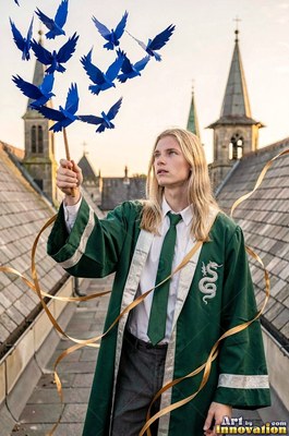 Standing on a castle roof, a blond man in a long green robe holds a stick of blue paper birds