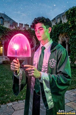 Dressed in a green robe in a castle courtyard, a young man cradles a wand under a bright pink shield