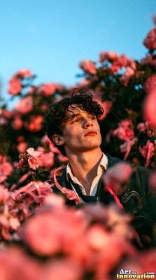 A young man stands outside behind pink rose bushes