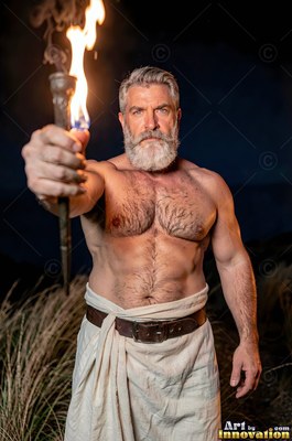 In the dark outdoors, an athletic man with a beard holds forward a burning torch