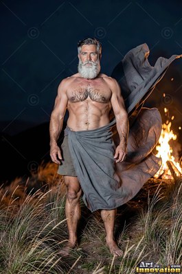 Bearded man in a blowing gray cloth standing by a fire at night