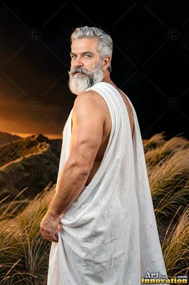 Glancing back over his bare shoulder, a gray-haired man with a beard wears a white robe on a dark hill