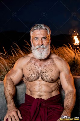 A shirtless, gray-haired man sporting a beard sits outside at night near warm torchlight