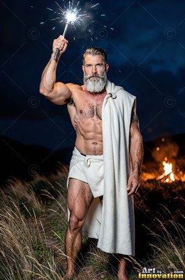 In a dark field, a well-built man who has a beard wrapped in a white robe holds up a sparkler
