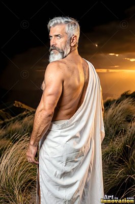 A white cloth draped over a muscular man standing in dry grass beneath a stormy sunset
