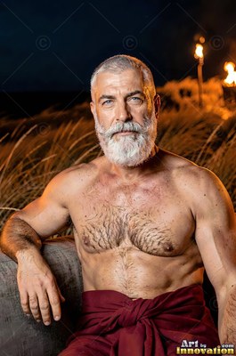 A shirtless older man with a white beard sits outside by tall grass at night