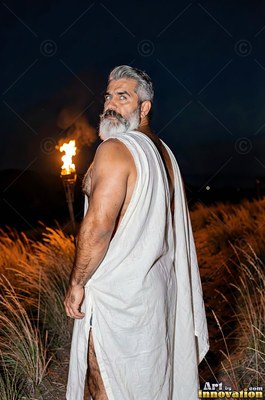 Wearing a white robe, a man sporting a beard looks back beside a torch in a dark field