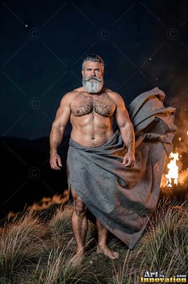 A muscular full-bearded man stands in dry grass at night wrapped in gray cloth