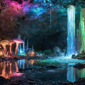 A vibrant and detailed fantasy photograph of an alien planet. Where every element appears meticulously crafted from iridescent stained glass. From a cave system, dominated by towering crystals, giant bioluminescent mushrooms, in all shapes and sizes, that glow in vibrant iridescent colors. A stream of iridescent blue water, flows down the center of the cave from a waterfall, off in the distance. The alien flora, vibrantly colored, attracting alien insects, like nothing you've ever seen. Glow worms hang down from the ceiling, emitting the entire spectrum of the rainbow in colors, they seem to dance and blend as if they are connected. Outside the cave system, the sky shimmers with a breathtaking aurora borealis that fills the sky, creating a cosmic spectacle of galaxies, nebulae, and radiant starlight, in this nighttime landscape.