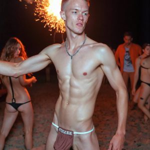 Bad Boys Naked Beach Party II. A full-body composition of a nude young man standing near a crackling bonfire, on a serene moonlit night at the beach. The gentle glow of the flames, flickers across his shinny wet body, catching his contours, and highlighting his muscular physique and handsome face. People everywhere partying, dancing to the music, having a good time together, naked, and free.