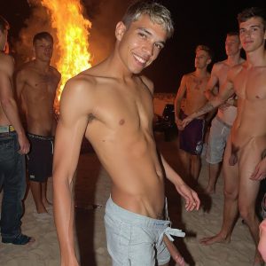 Bad Boys Naked Beach Party II. A full-body composition of a nude young man standing near a crackling bonfire, on a serene moonlit night at the beach. The gentle glow of the flames, flickers across his shinny wet body, catching his contours, and highlighting his muscular physique and handsome face. People everywhere partying, dancing to the music, having a good time together, naked, and free.