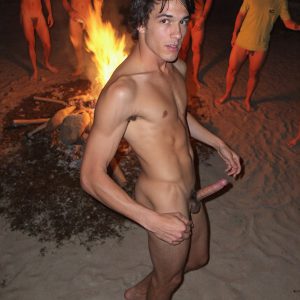 Bad Boys Naked Beach Party II. A full-body composition of a nude young man standing near a crackling bonfire, on a serene moonlit night at the beach. The gentle glow of the flames, flickers across his shinny wet body, catching his contours, and highlighting his muscular physique and handsome face. People everywhere partying, dancing to the music, having a good time together, naked, and free.