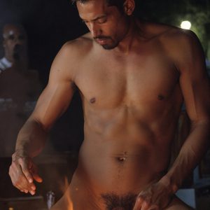 Bad Boys Naked Beach Party II. A full-body composition of a nude young man standing near a crackling bonfire, on a serene moonlit night at the beach. The gentle glow of the flames, flickers across his shinny wet body, catching his contours, and highlighting his muscular physique and handsome face. People everywhere partying, dancing to the music, having a good time together, naked, and free.