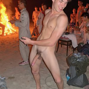 Bad Boys Naked Beach Party II. A full-body composition of a nude young man standing near a crackling bonfire, on a serene moonlit night at the beach. The gentle glow of the flames, flickers across his shinny wet body, catching his contours, and highlighting his muscular physique and handsome face. People everywhere partying, dancing to the music, having a good time together, naked, and free.