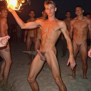 Bad Boys Naked Beach Party II. A full-body composition of a nude young man standing near a crackling bonfire, on a serene moonlit night at the beach. The gentle glow of the flames, flickers across his shinny wet body, catching his contours, and highlighting his muscular physique and handsome face. People everywhere partying, dancing to the music, having a good time together, naked, and free.
