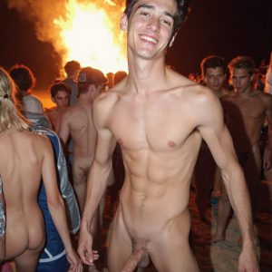 Bad Boys Naked Beach Party II. A full-body composition of a nude young man standing near a crackling bonfire, on a serene moonlit night at the beach. The gentle glow of the flames, flickers across his shinny wet body, catching his contours, and highlighting his muscular physique and handsome face. People everywhere partying, dancing to the music, having a good time together, naked, and free.